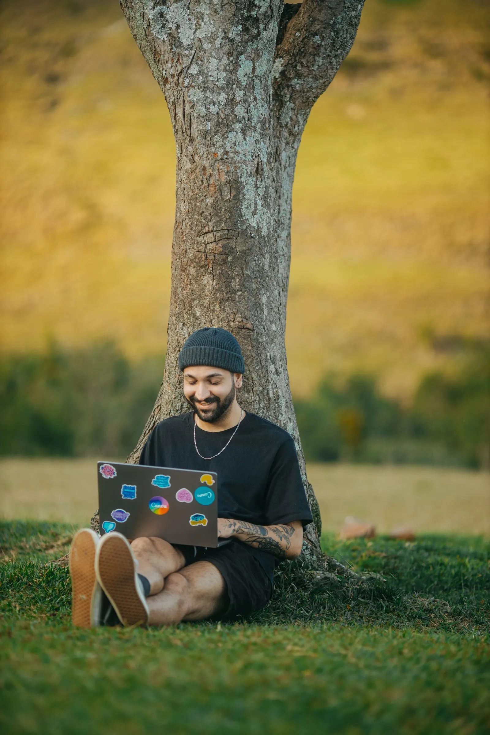 Ein Mann sitzt mit Laptop unter einem Baum auf der Wiese und lacht.